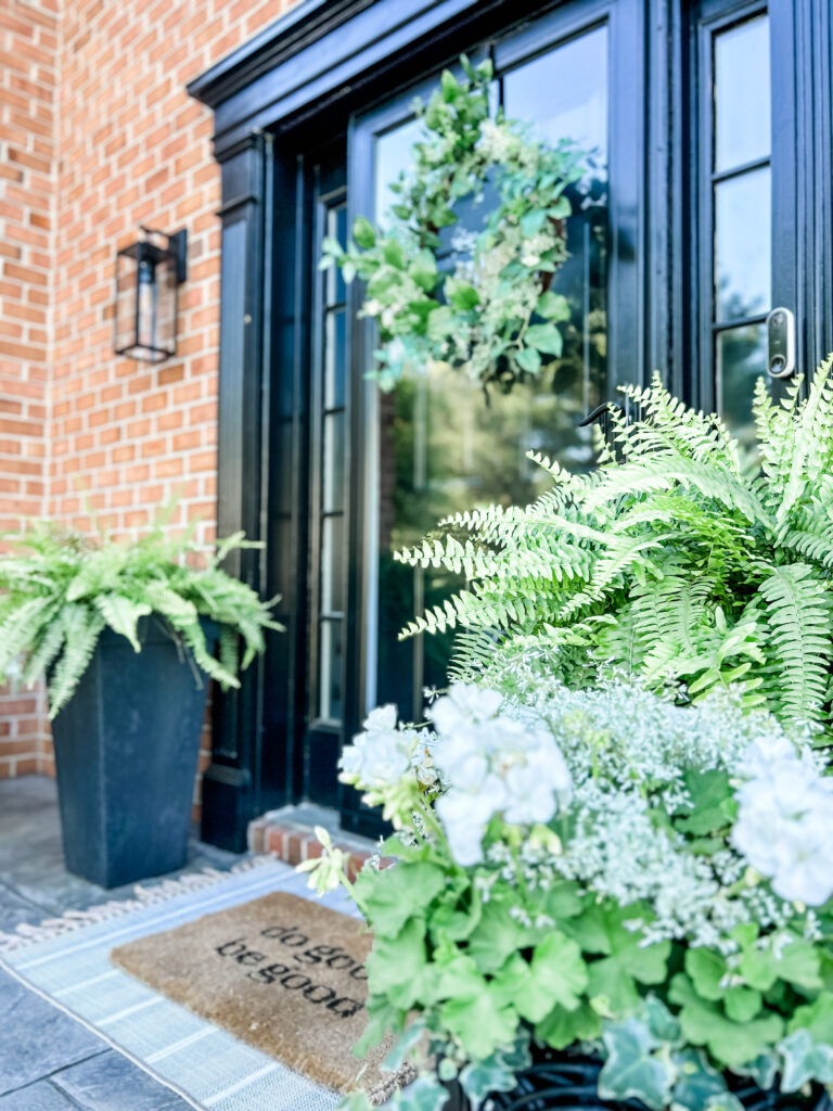 Welcoming Front Porch front entryway featuring black planters, a lush green wreath, and a layered "do good be good" doormat